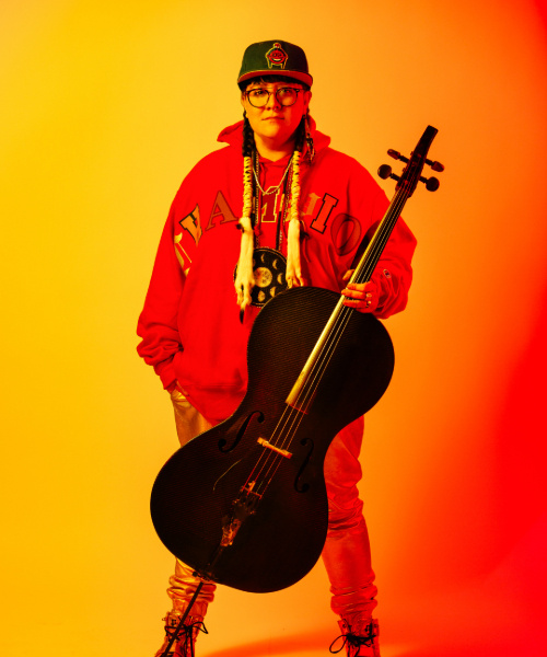 A performer stands with a large stringed instrument in front of a yellow-orange gradient.